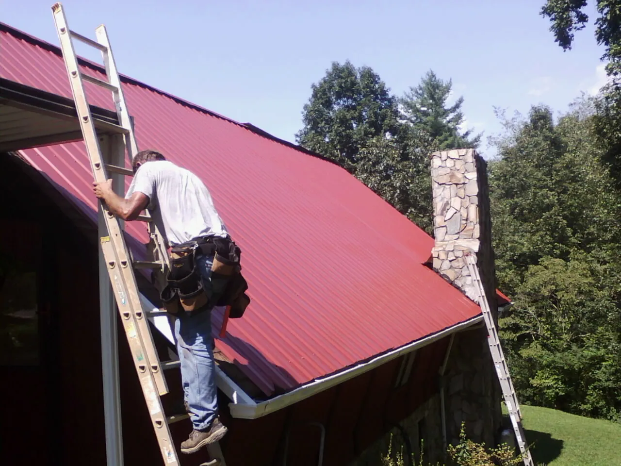 Roofer on a ladder beside a red metal roof and stone chimney — Soffit & Fascia Repair by Roofing Brothers in Bellingham